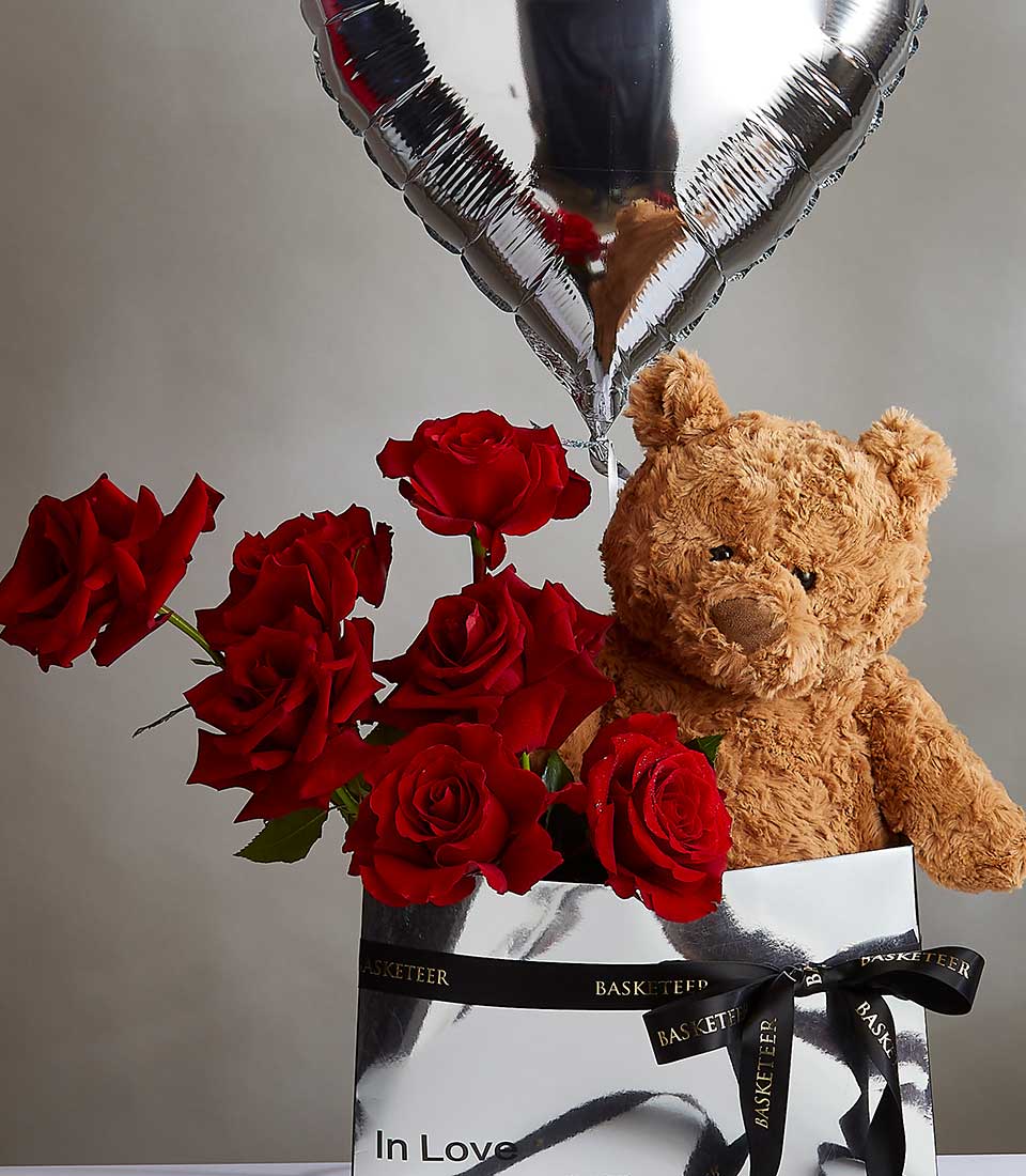 A silver box with black ribbons holds a heartfelt bouquet of red roses, a brown teddy bear, and a silver heart-shaped balloon. The teddy bear is nestled among the roses, and the box features the words 