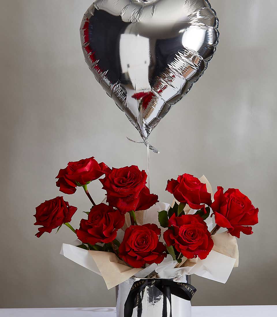 A silver heart-shaped balloon floats above a Passionate Rose Heart Bouquet, with vibrant red roses arranged in a white vase wrapped with a black ribbon. The soft gray background enhances the vivid color of the roses and the reflective shine of the balloon.