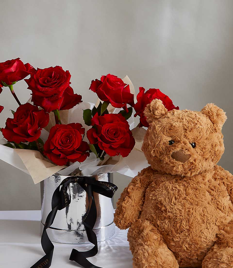 A bouquet of red roses wrapped in white paper is displayed in a shiny silver vase with a black ribbon tied around it. Next to the vase is a fluffy, brown teddy bear labeled as the Ruby Rose Teddy Surprise. Both items are set against a neutral background.