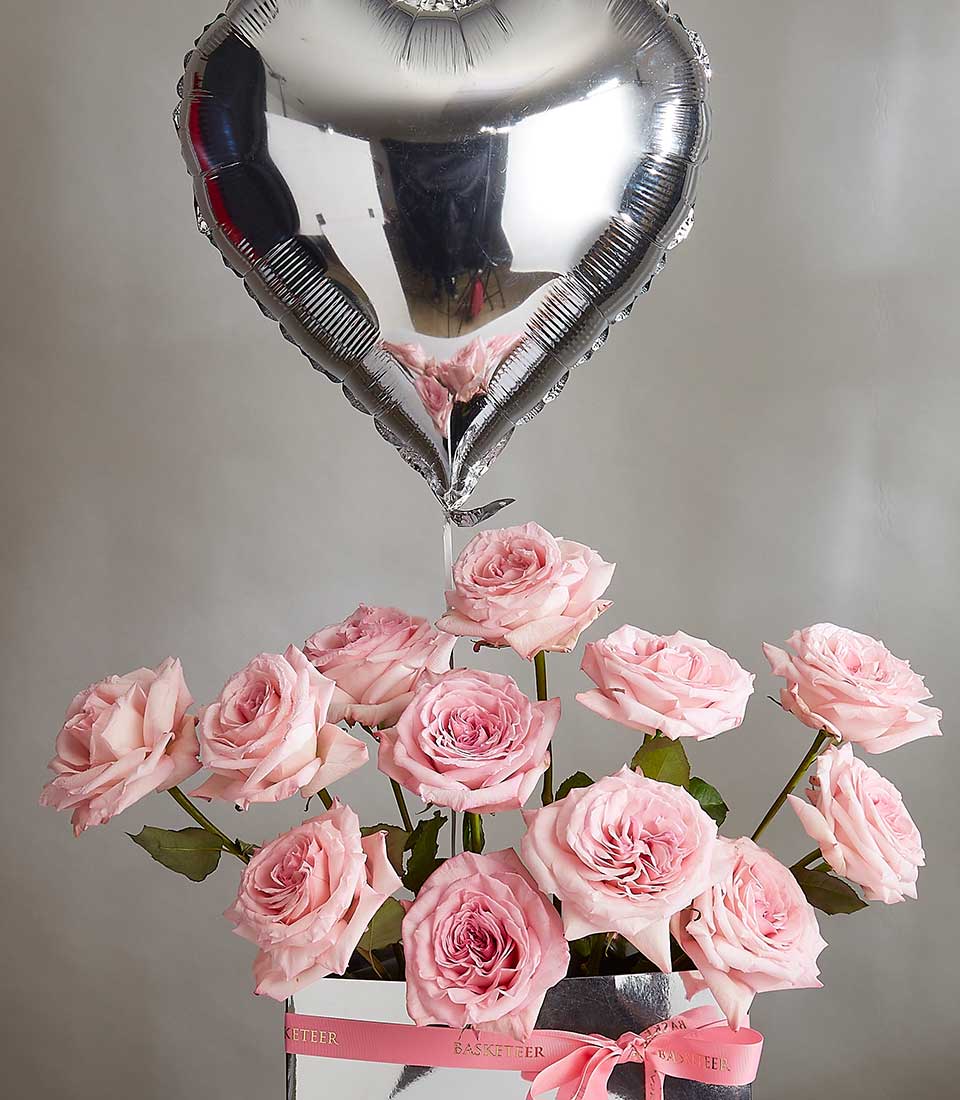 A heartfelt O'Hara surprise package features a bouquet of light pink roses elegantly arranged in a white box with a pink ribbon. Above the bouquet, a large silver heart-shaped balloon floats, tethered to the box. The background is a neutral gray.