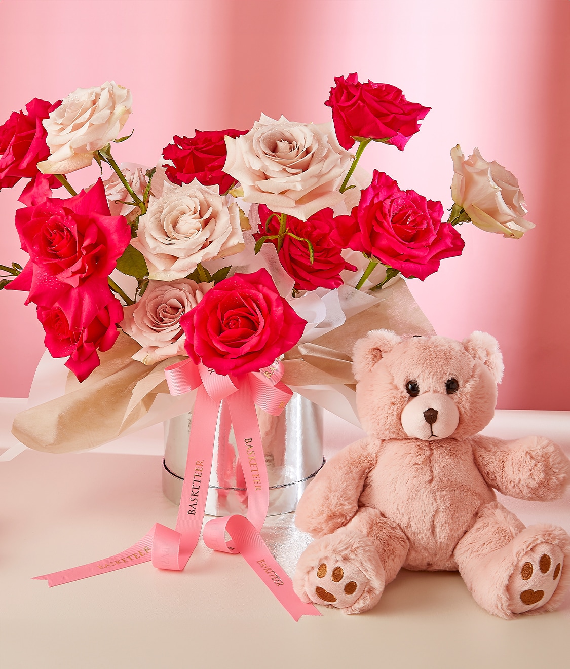 A clear vase holds a bouquet of pink and white roses, adorned with pink ribbons, situated on a light pink surface. Beside the vase is a small light brown teddy bear and a single pink rose, against a soft pink background.