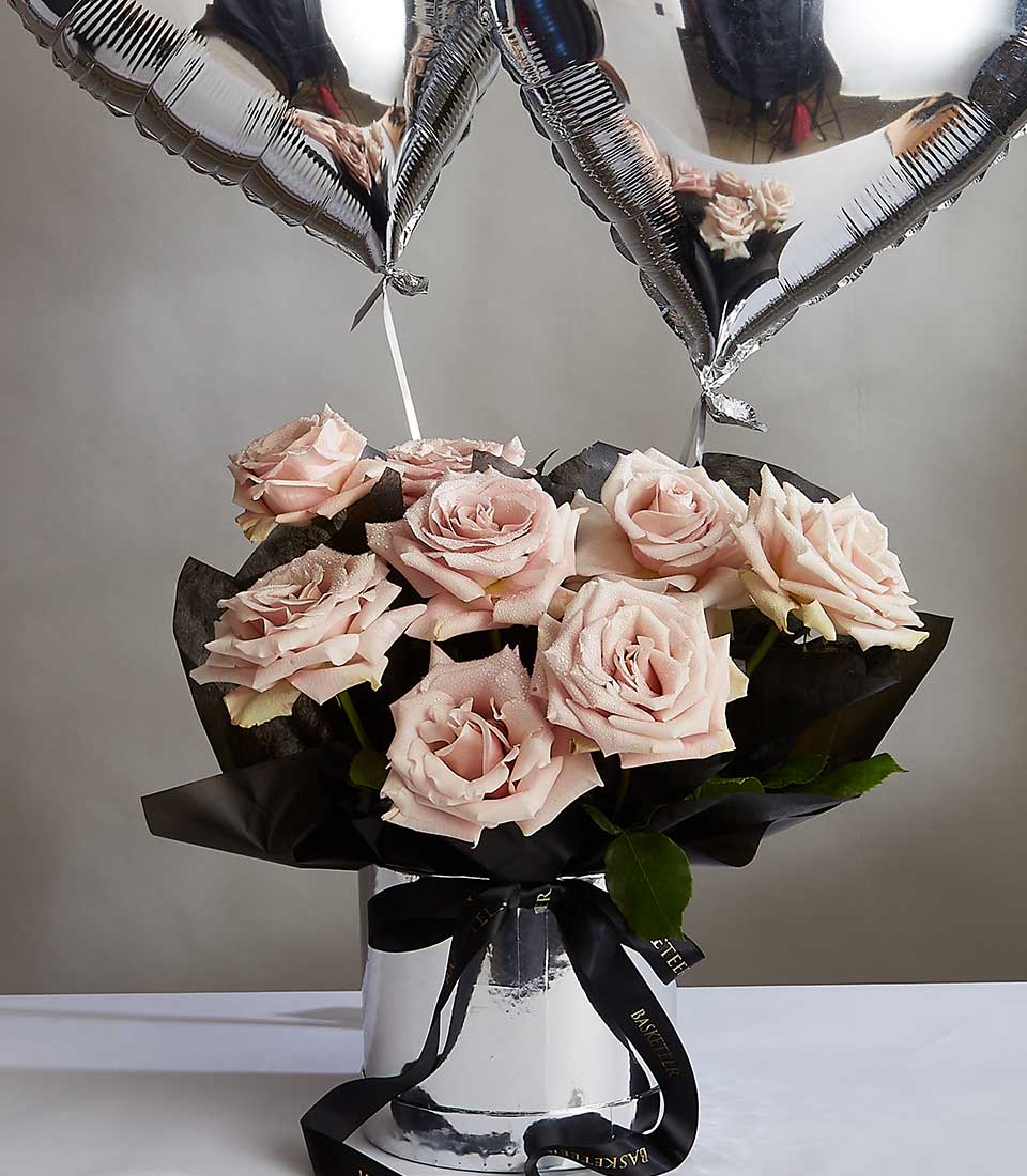 The Rose Radiance Gift Bundle features a bouquet of blush pink roses arranged in a reflective silver vase, accompanied by two metallic silver heart-shaped balloons. The vase is adorned with a black ribbon, and the background showcases a neutral gray wall.