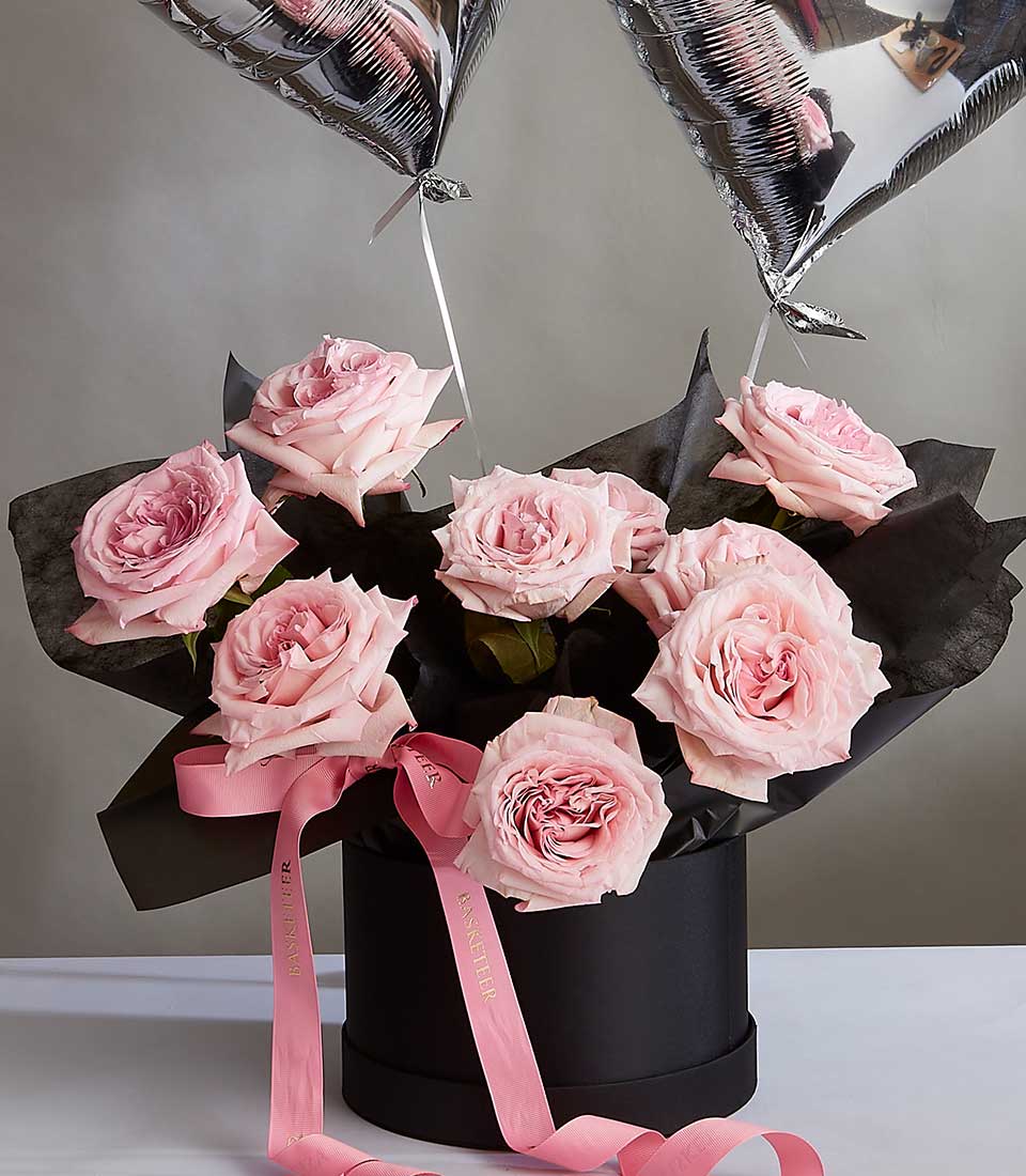 A bouquet of large, Pink O'Hara roses arranged in a black cylindrical vase with pink ribbons and metallic silver heart-shaped balloons attached, placed on a white surface against a neutral background. The roses are wrapped in black tissue paper within the vase.