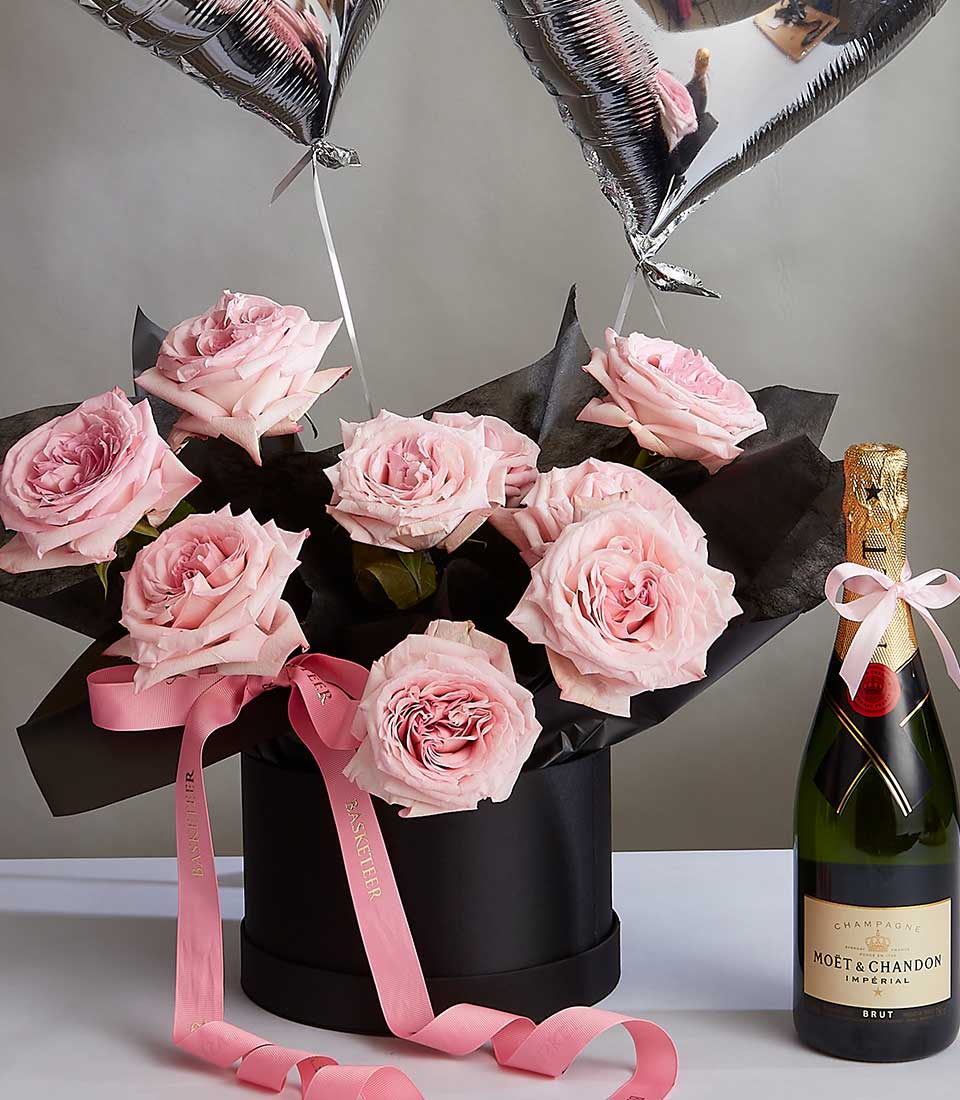 A round black box from the Blush Rose Serenade Collection holds pink roses with two silver heart-shaped balloons attached. A pink ribbon with text is draped over the box. To the right, a bottle of Moët & Chandon champagne, adorned with a pink ribbon bow, stands upright.