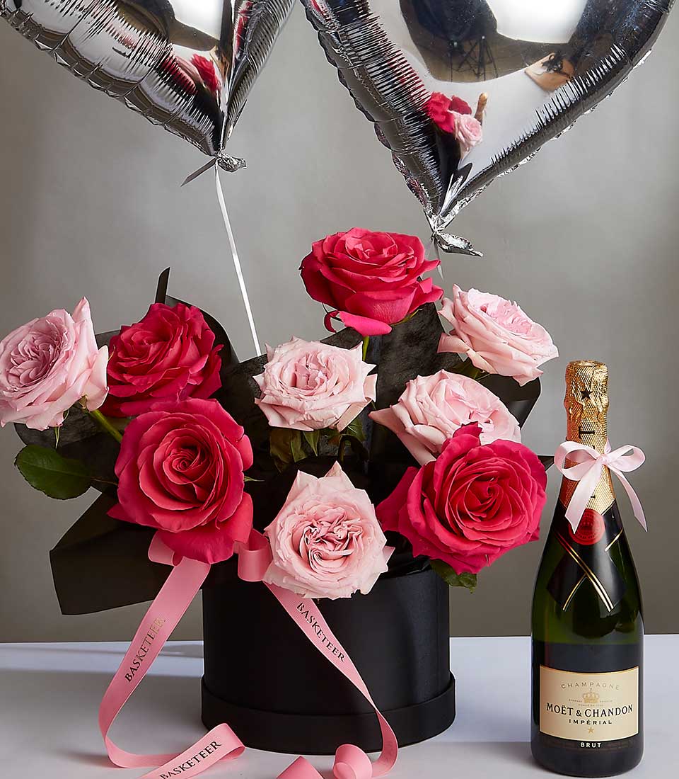 A black hatbox filled with vibrant pink and blush roses is adorned with two heart-shaped silver balloons. Pink ribbons labeled 