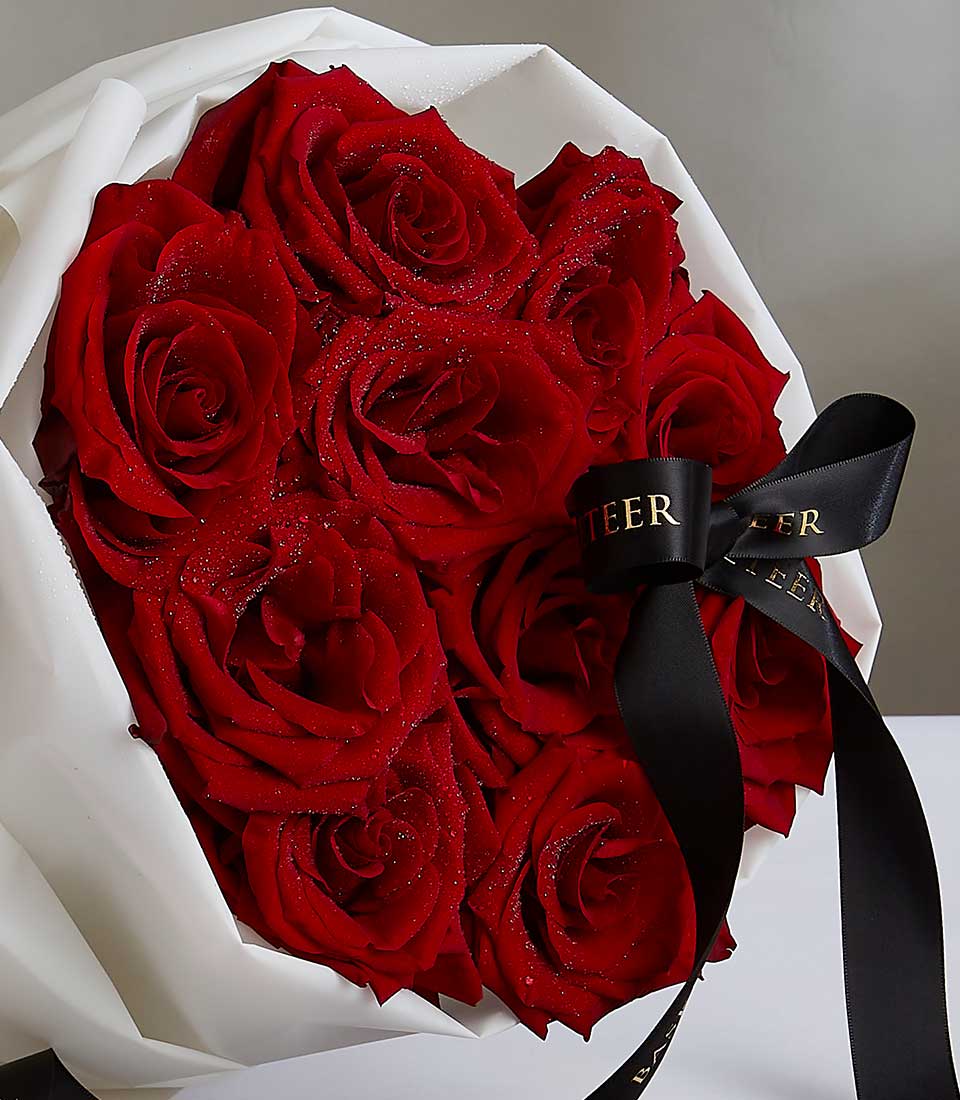 A bouquet of vibrant Red Explorer Roses wrapped in white paper, adorned with a black ribbon featuring gold writing. The roses are fresh and appear velvety with a sprinkle of glitter, presenting a luxurious and elegant arrangement.
