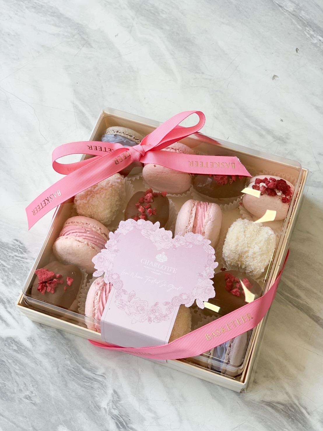 A luxury macaron and chocolate gift box wrapped with a pink Basketeer ribbon, featuring handcrafted sweets in shades of pink and white.