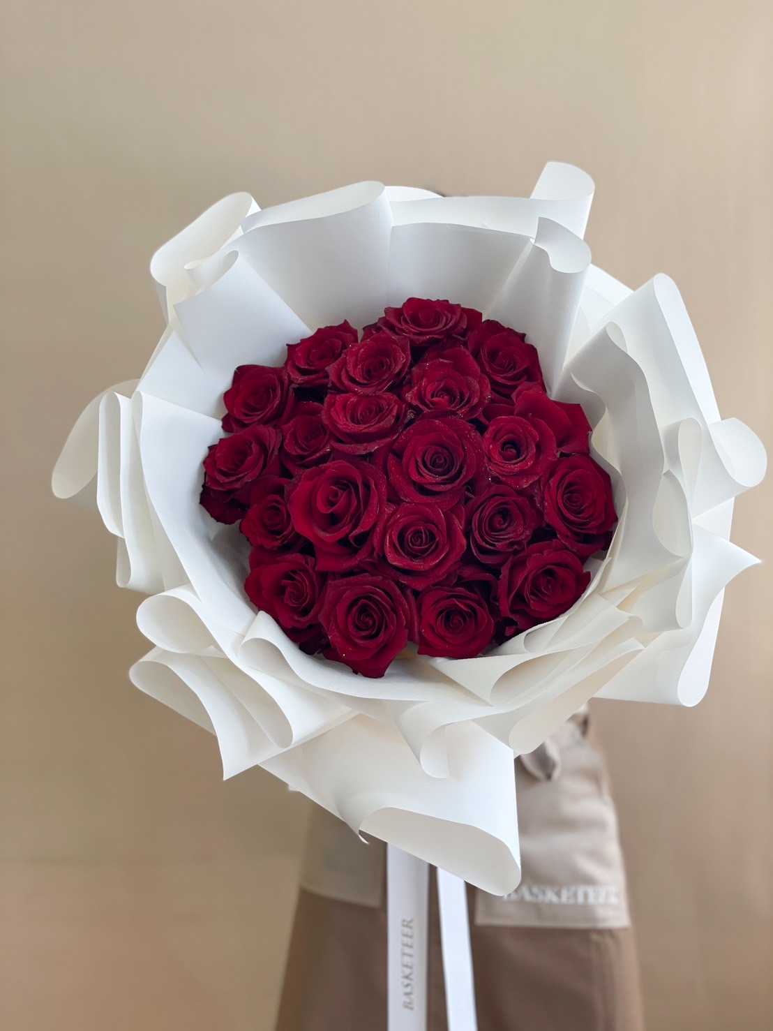 A luxurious hand-tied bouquet of premium red roses, elegantly wrapped in white paper with a Basketeer ribbon.