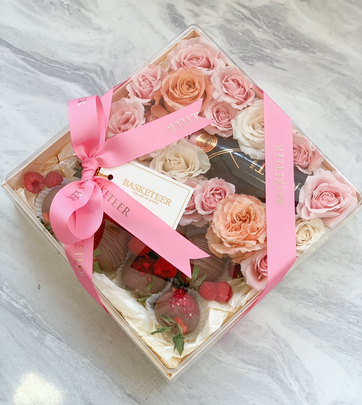 Luxury champagne and chocolate-covered strawberry gift box with preserved roses, elegantly wrapped in a transparent case with a pink Basketeer ribbon.