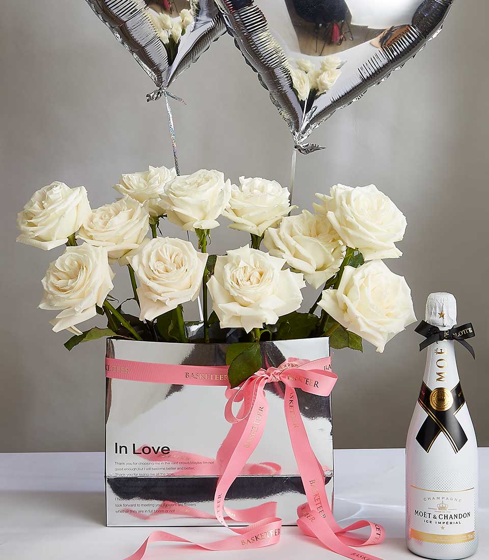 A gift set featuring a bouquet of white roses in a rectangular silver vase with pink ribbon, two silver heart-shaped balloons, and a bottle of Moët & Chandon champagne adorned with a black bow. The vase has the words 