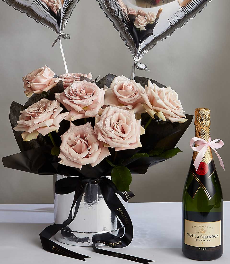 A bouquet of light pink roses arranged in a silver cylindrical vase wrapped with black ribbon, paired with a bottle of Moët & Chandon champagne. Two silver, heart-shaped balloons are attached to the bouquet, creating an elegant celebratory display.