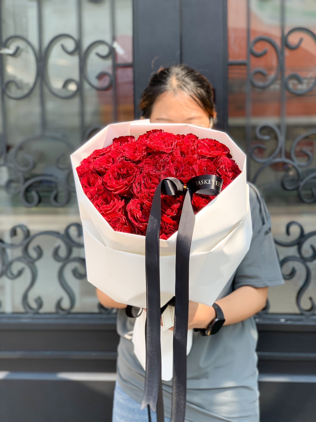 A grand bouquet of luxurious deep red roses, wrapped in white paper with a black ribbon, held by a person in front of a decorative gate.