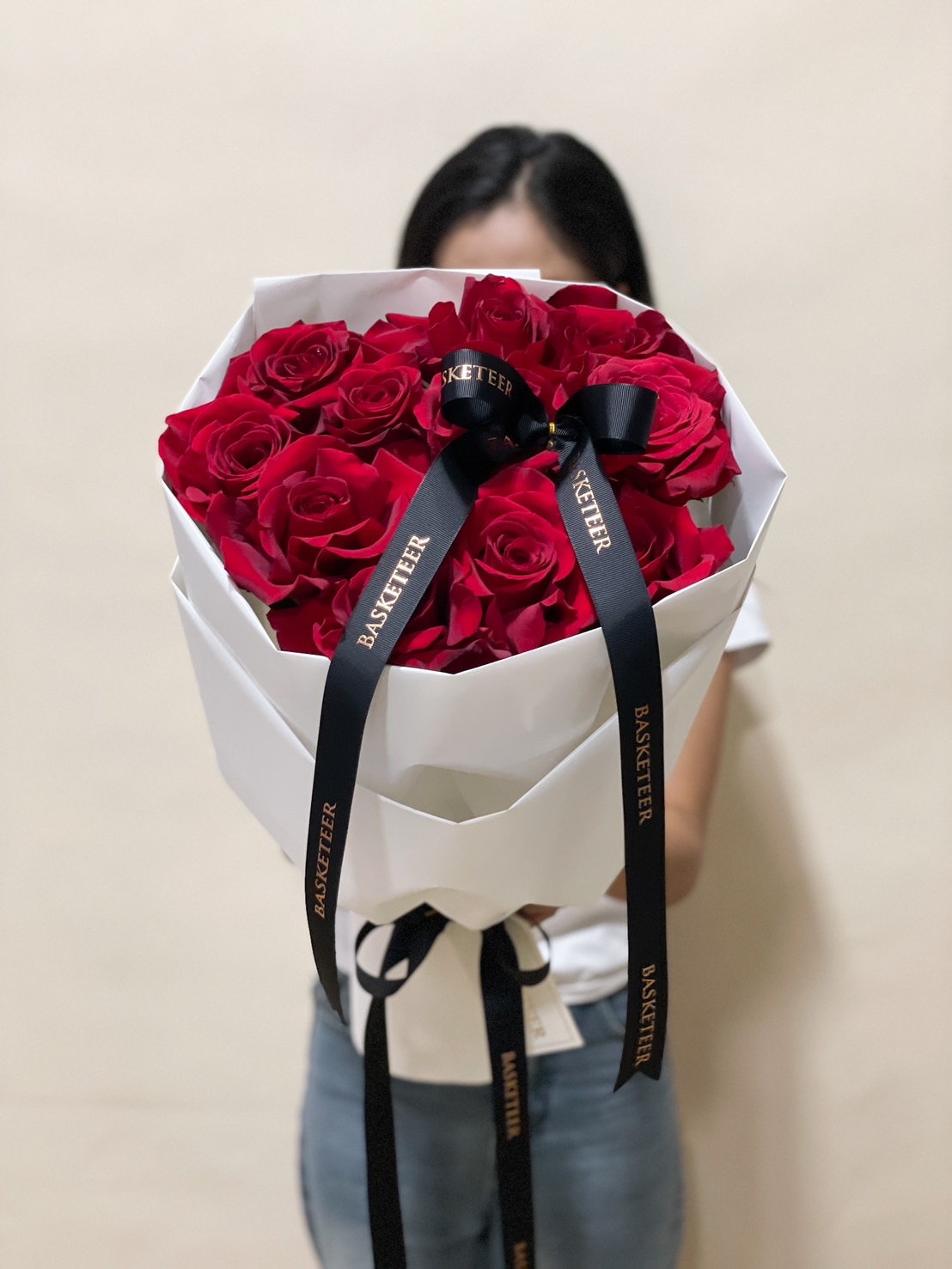 A grand bouquet of luxurious deep red roses, wrapped in white paper with a black ribbon, held by a person in front of a decorative gate.