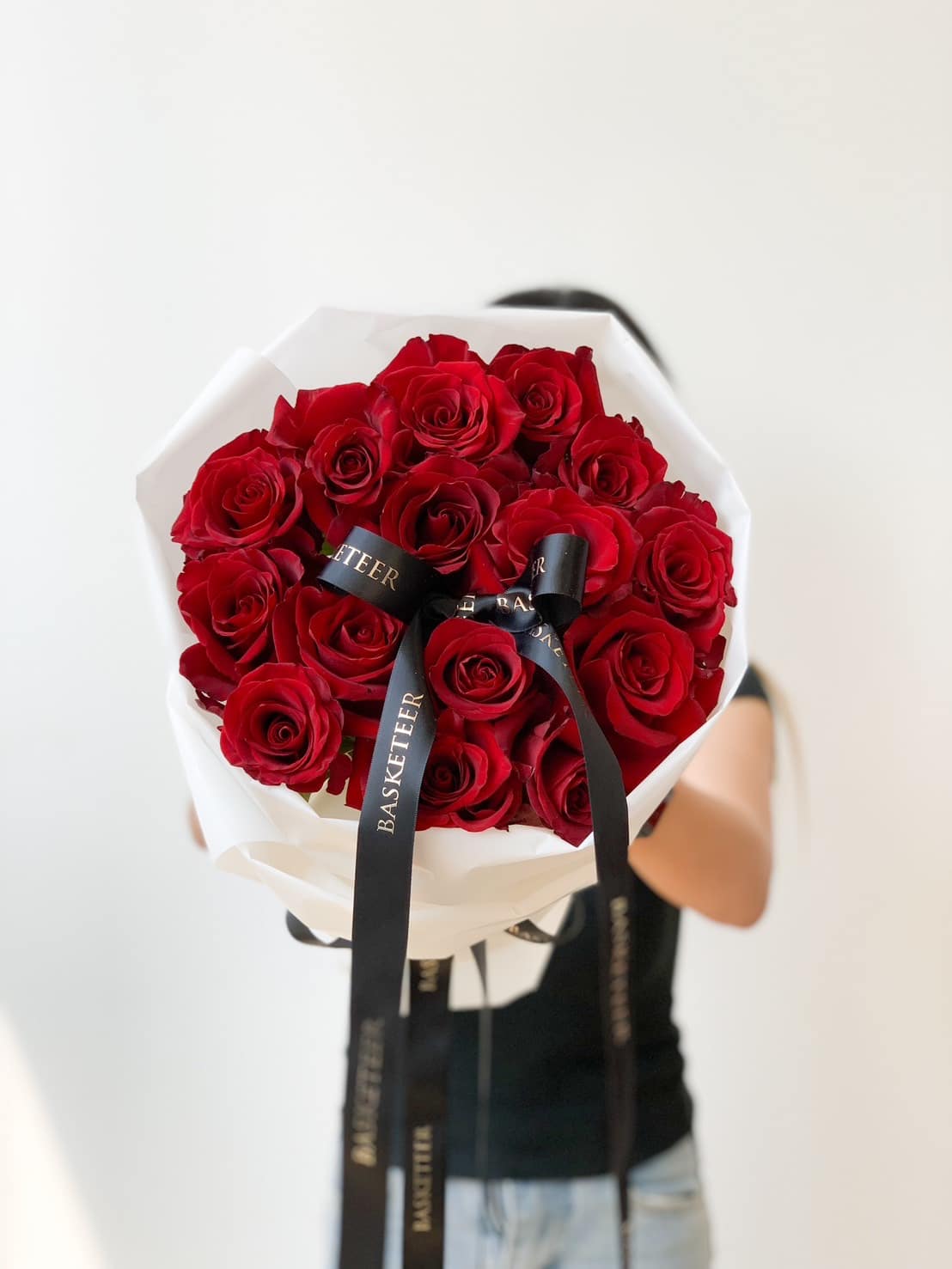 A person holding a Red Explorer Roses Bouquet wrapped in white paper, adorned with black ribbons that have 