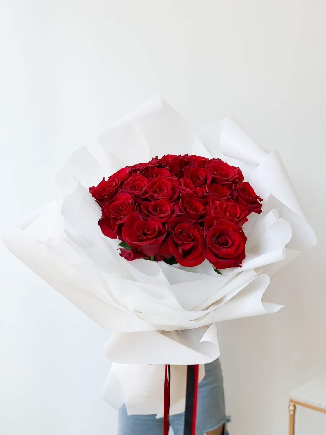 A large Explorer Red Roses Bouquet is beautifully wrapped in layers of white paper. The vibrant red roses are held in front of a plain white background, partially obscuring the person holding it.
