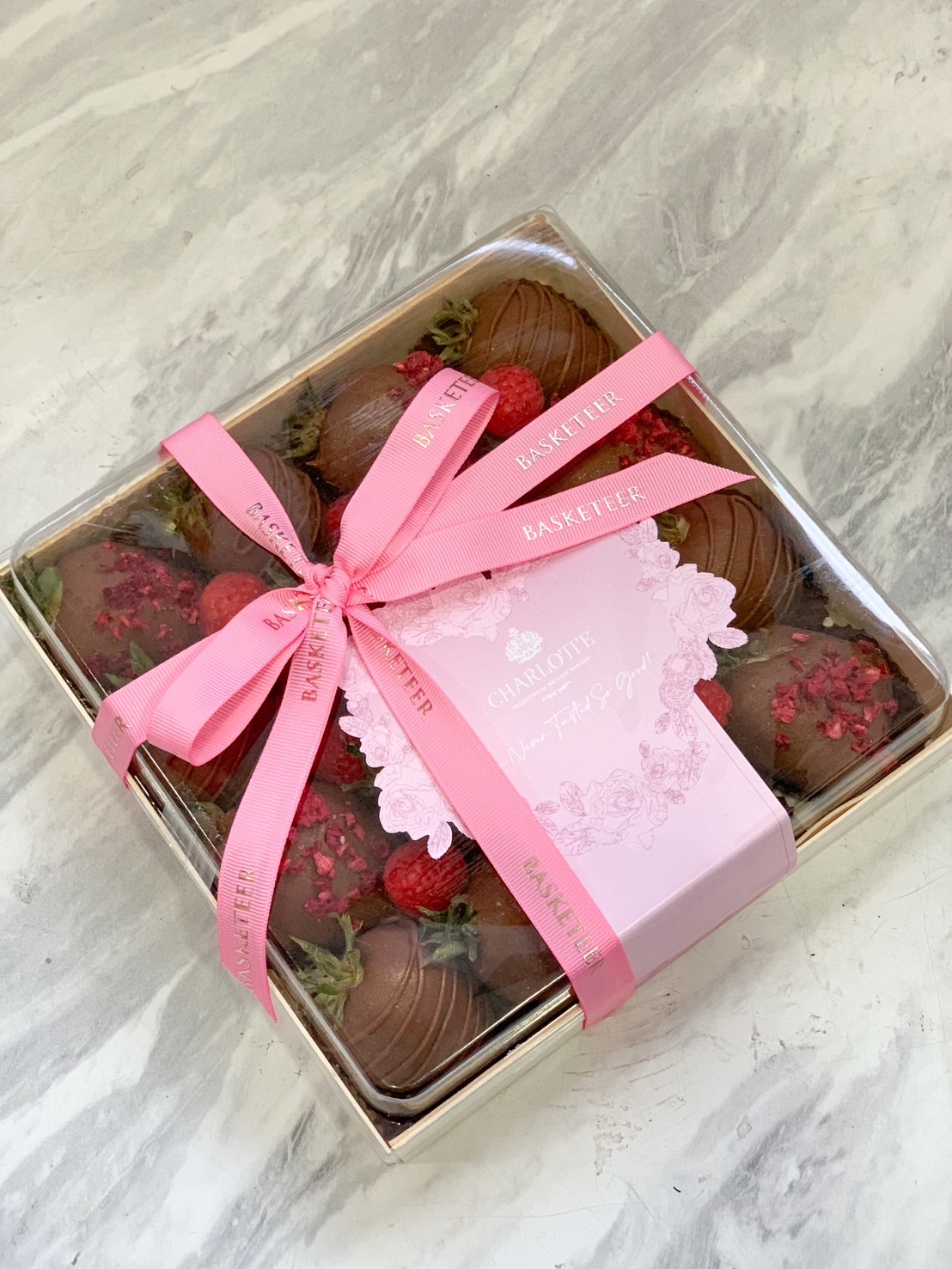 A beautifully wrapped gift box of chocolate-covered strawberries, adorned with a pink satin ribbon, featuring fresh raspberries and elegant packaging by Basketeer.