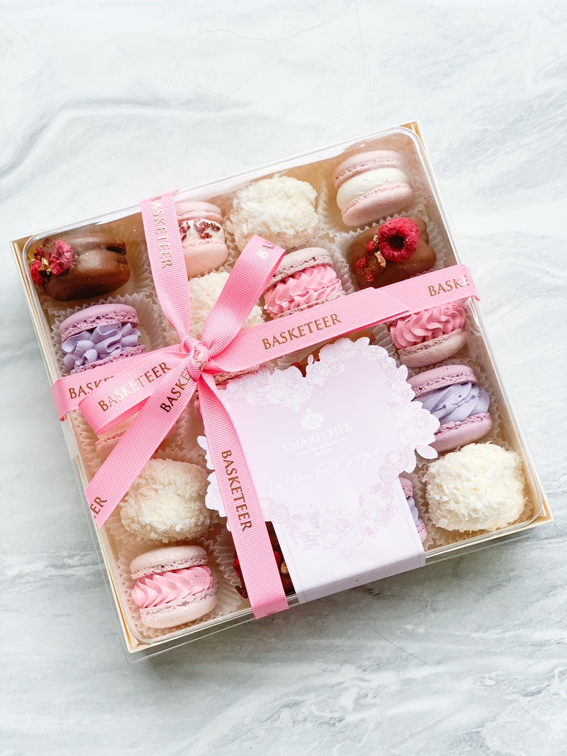 A luxurious gift box, featuring an exquisite assortment of pastel-colored macarons and delicately coconut-covered treats, is elegantly tied with a pink ribbon showcasing the 