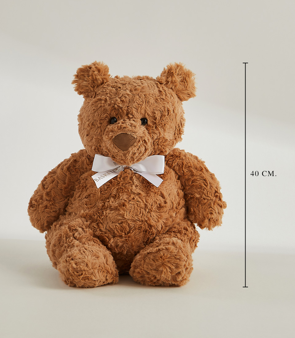 A large, fluffy teddy bear holds a wooden fork in its right paw and has cake crumbs around its mouth. It is seated at a table adorned with a small slice of yellow cake with pink frosting on a white plate. Beside the bear, a White Playa Blanca Roses Bouquet adds an elegant touch. The background is plain gray.