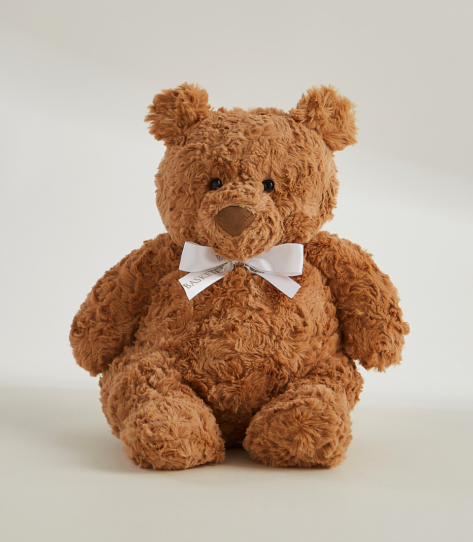 A large, fluffy teddy bear holds a wooden fork in its right paw and has cake crumbs around its mouth. It is seated at a table adorned with a small slice of yellow cake with pink frosting on a white plate. Beside the bear, a White Playa Blanca Roses Bouquet adds an elegant touch. The background is plain gray.