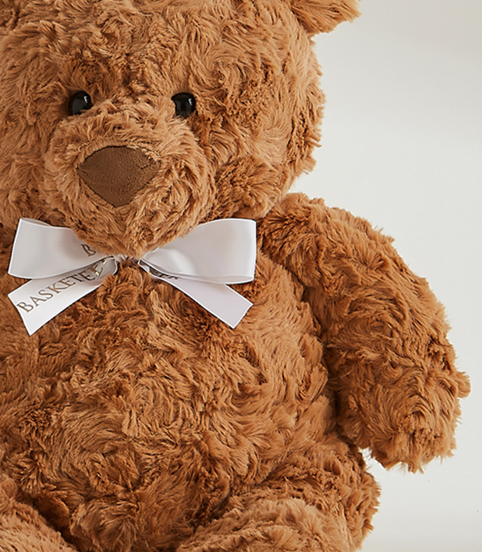 A large, fluffy teddy bear holds a wooden fork in its right paw and has cake crumbs around its mouth. It is seated at a table adorned with a small slice of yellow cake with pink frosting on a white plate. Beside the bear, a White Playa Blanca Roses Bouquet adds an elegant touch. The background is plain gray.