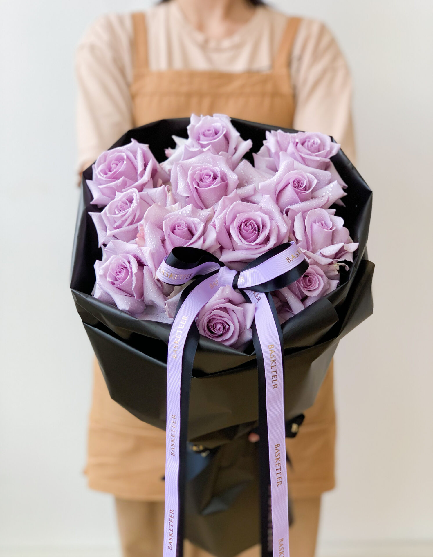 Bouquet of lavender roses wrapped in black paper with purple ribbons, held by a person in a beige apron.