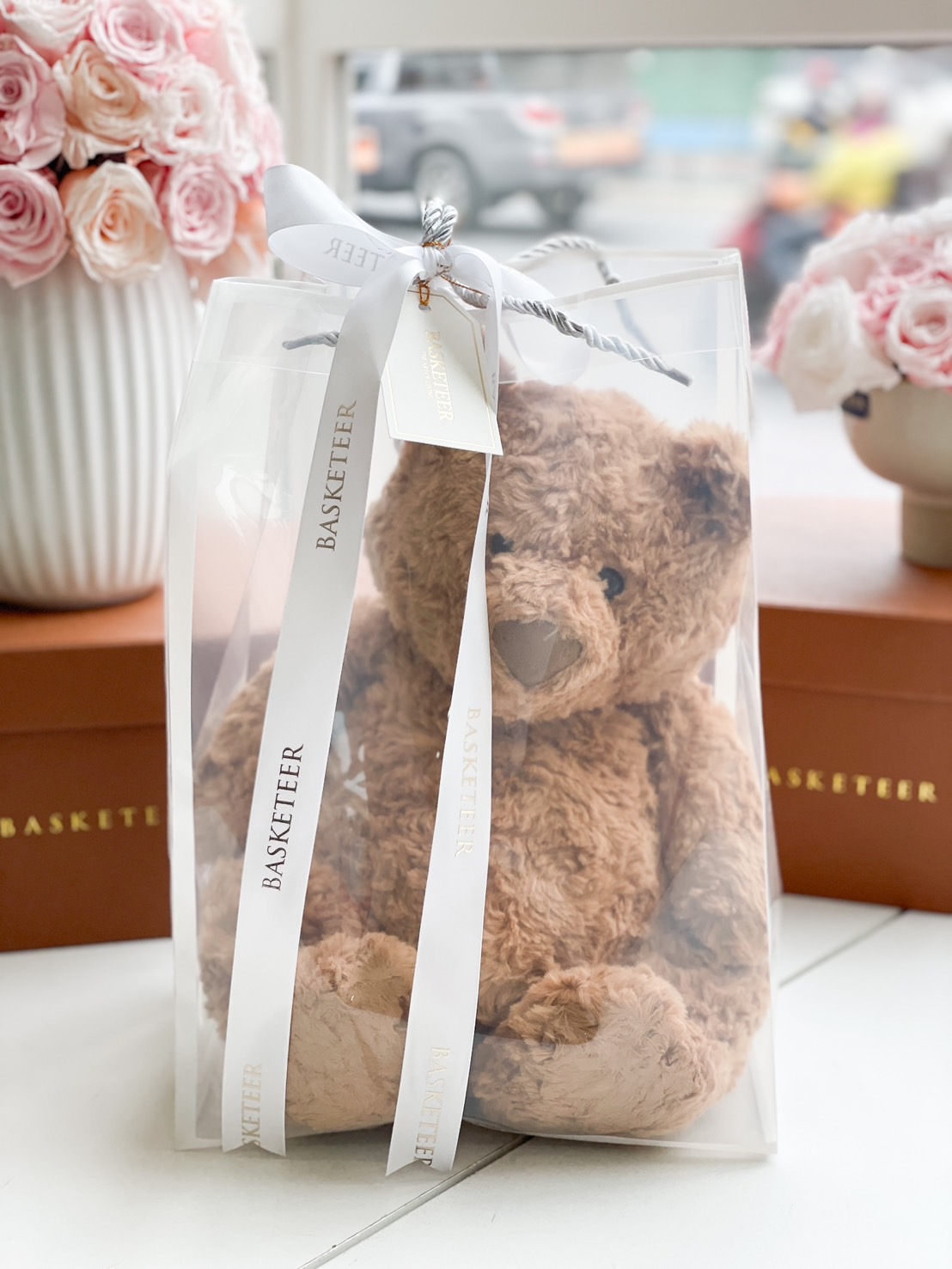 A soft brown teddy bear elegantly packaged in a transparent gift bag, tied with a white 