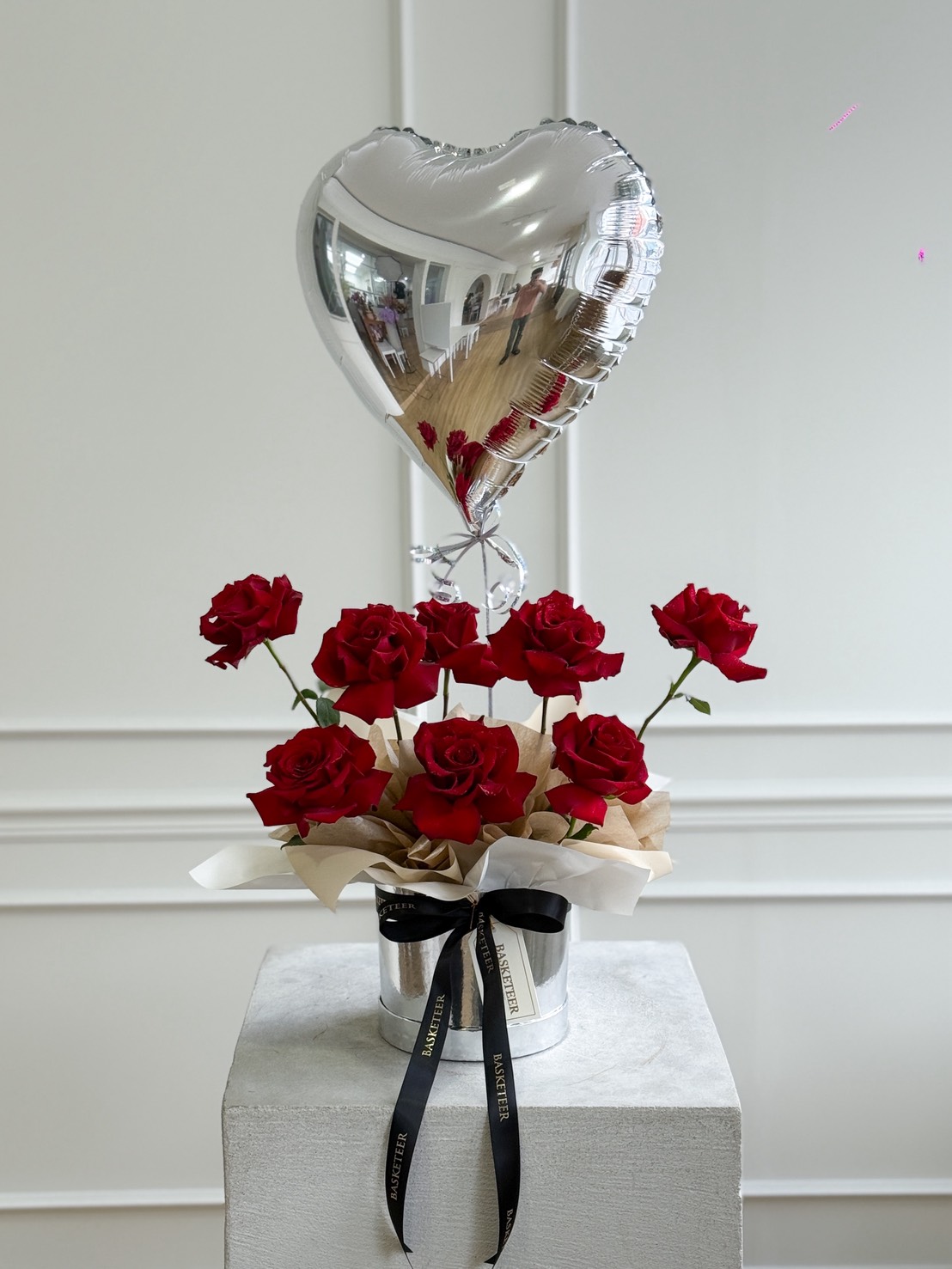 The Passionate Rose Heart Bouquet features red roses in a white hat box with a black ribbon, crowned by a shiny silver heart-shaped balloon and elegantly displayed on a pedestal against a white wall.