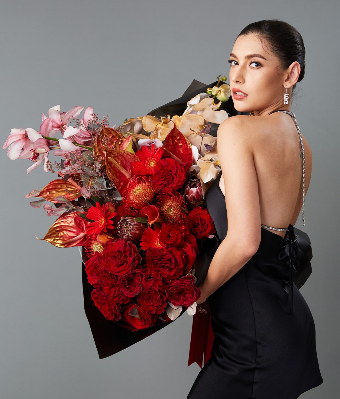A woman with dark hair styled in an elegant updo, wearing a backless black dress and dangling earrings, holds a large, vibrant bouquet of red, pink, and cream flowers, posing against a plain gray background. Her expression is poised and confident.