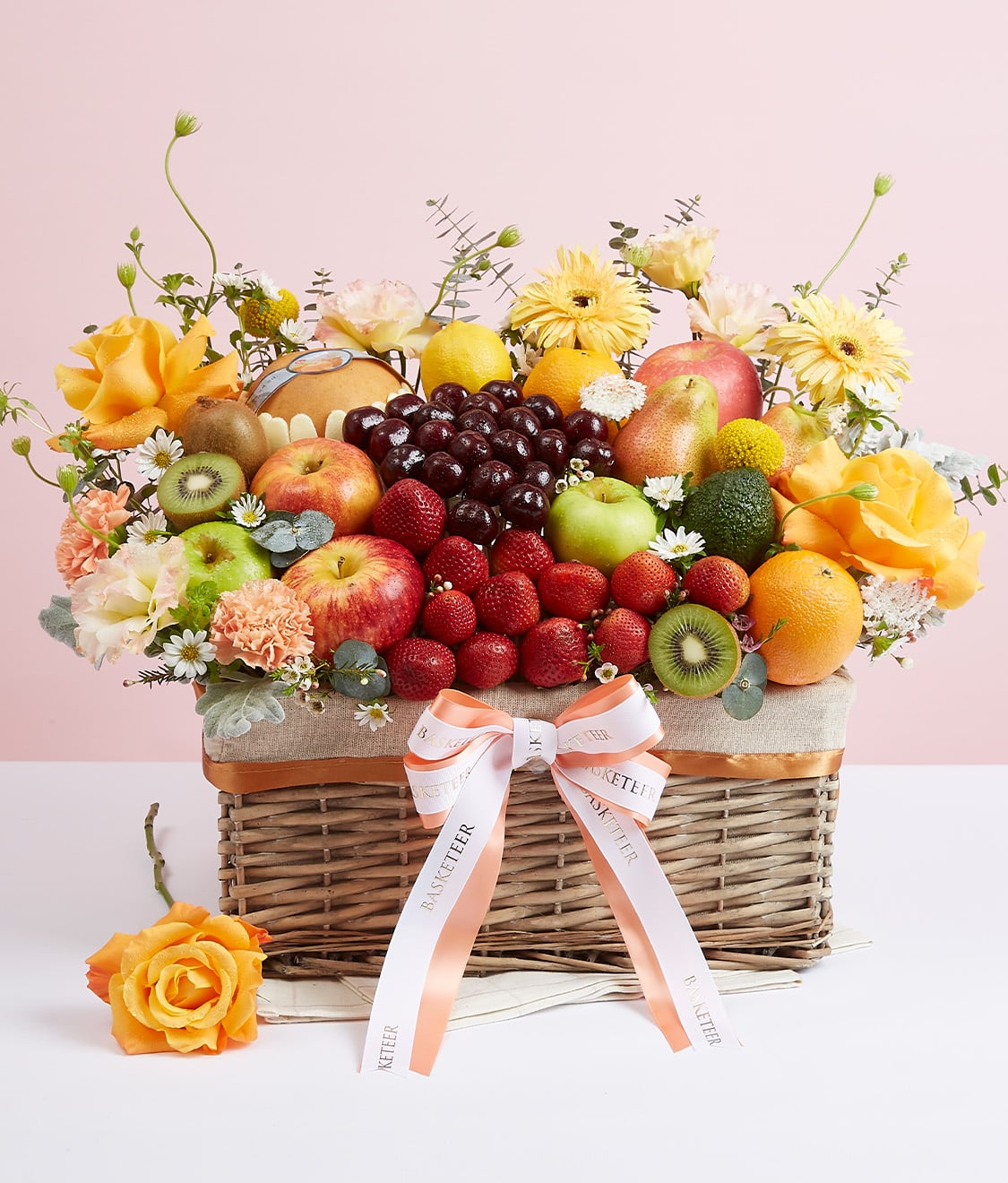A woven basket filled with an assortment of fresh fruits, including apples, oranges, grapes, and strawberries, decorated with yellow and pink flowers. The basket has a white ribbon tied in a bow on the front, and a single yellow rose lies beside it on a white surface.