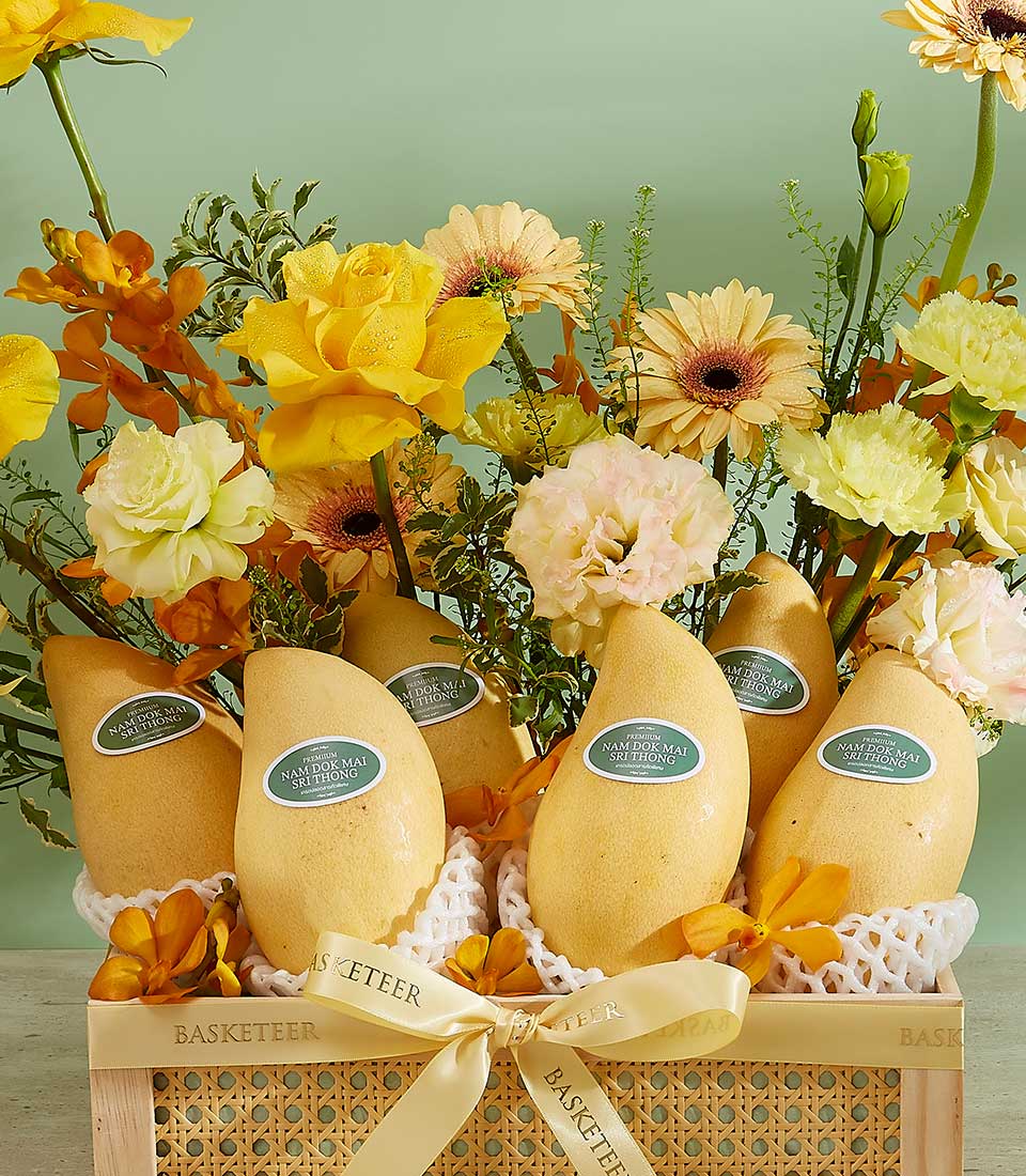 A woven basket filled with yellow and orange flowers and several large mangoes. Each mango has a protective foam covering at the bottom and a green sticker on it. A yellow ribbon with the words 