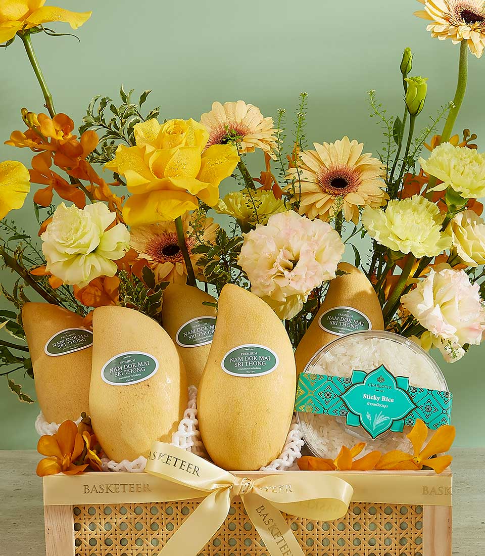 A decorative basket showcases six Nam Dok Mai Thai mangoes with labels, surrounded by various yellow and white flowers. To the right of the mangoes, there's a clear container of Splendor Rice Delight. The basket is adorned with a ribbon labeled 