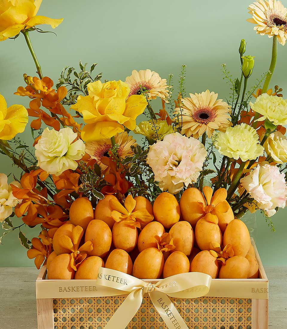 A premium Mayongchid Blissful Hamper features a wicker basket filled with a variety of fresh yellow and cream flowers, including roses, gerbera daisies, and orchids. The front section of the basket holds numerous ripe oranges, all tied with orange ribbons.