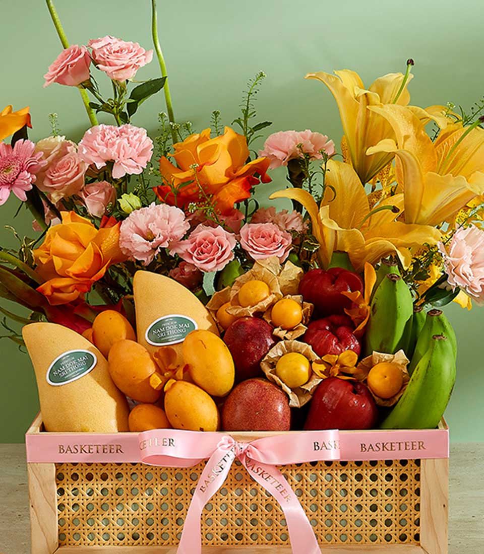 A decorative wooden basket filled with an assortment of fruits including mangoes, bananas, and apples, surrounded by vibrant flowers like lilies, roses, and carnations. This Tropical Harvest Gift Basket is adorned with a pink ribbon featuring the name 