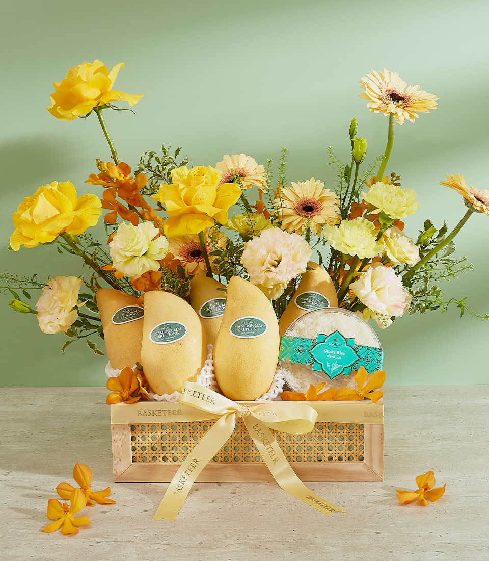 A decorative wooden basket is filled with four large yellow Nam Dok Mai mangoes and an assortment of vibrant yellow and white flowers. The basket is adorned with yellow ribbons and a small turquoise gift box, arranged against a pastel green background.