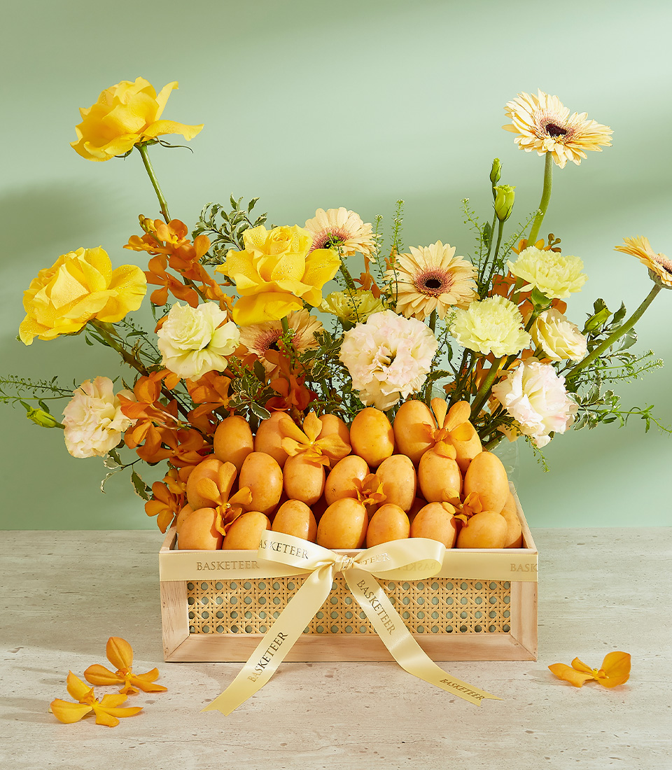 A wooden rectangular basket filled with fresh loquats and wrapped in a cream-colored ribbon with gold 