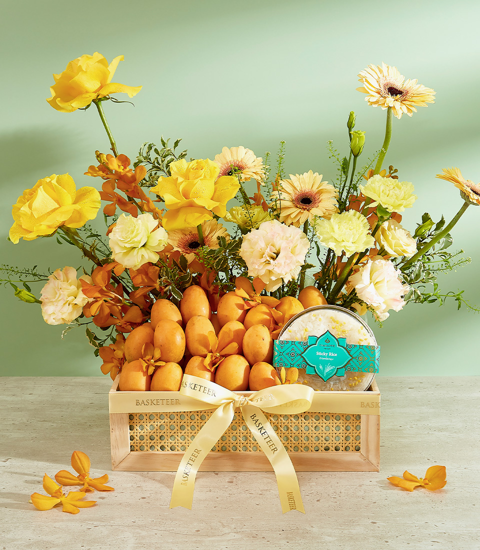A wooden crate containing yellow and orange flowers, such as roses and gerberas, arranged with ripe dates. The crate boasts a yellow ribbon and a round, turquoise tin with Arabic writing. This Luxe Mayongchid Gift Basket is set against a light green background.