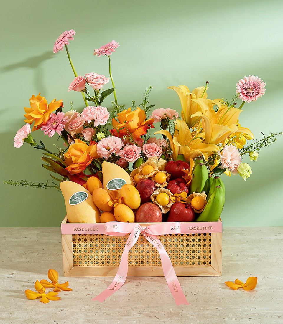 A Tropical Harvest Gift Basket filled with a vibrant assortment of fruits and fresh flowers. The fruits include mangoes, apples, pears, grapes, bananas, and oranges. The flowers are a mix of pink carnations, yellow lilies, pink gerbera daisies, and green foliage. The basket is adorned with a pink ribbon.