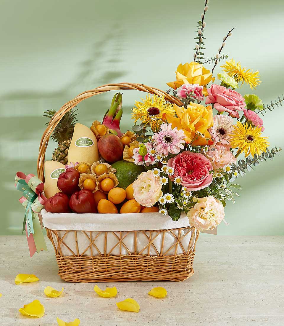 A vibrant tropical fruits basket brimming with a colorful assortment of fruits and flowers. The fruits include melons, lychees, apples, and dragon fruit. The flowers feature yellow roses, pink carnations, and various daisies, complemented by greenery. Yellow petals are scattered on the table.