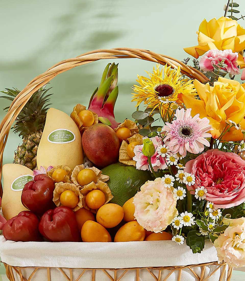 A woven basket brimming with a colorful assortment of vibrant tropical fruits and flowers. The fruits include mangoes, dragon fruit, grapes, star apples, and Physalis. The basket is adorned with vibrant yellow roses, pink daisies, sunflowers, and white blossoms.
