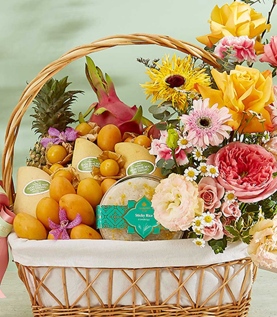 An exquisite Thai fruits hamper featuring a wicker basket filled with an assortment of tropical delights like mangos and a pineapple. Adorned with vibrant flowers in various colors, this cheerful arrangement brings a touch of paradise to any setting.