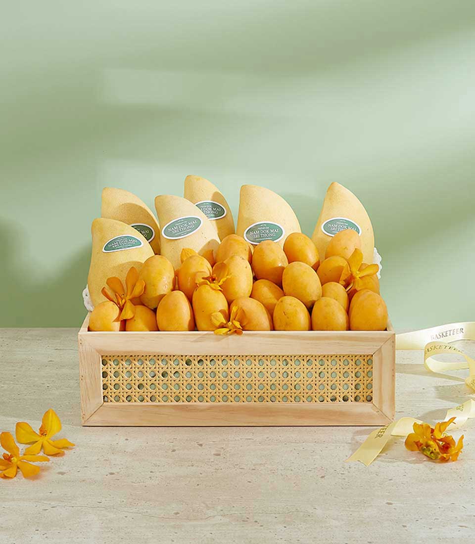 A wooden basket filled with premium summer fruits, including yellow mangoes adorned with yellow flowers, is placed on a light-colored table against a green background. Some mangoes have labels, and a yellow ribbon lies beside the basket.