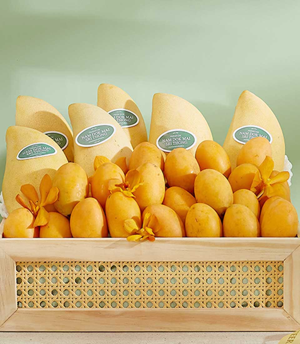 A rectangular, woven-pattern basket filled with premium summer fruits is displayed. The back row showcases larger, oblong Nam Dok Mai mangoes with labels, while the front row is adorned with smaller, rounder fruits—possibly plums—accentuated by small yellow flowers.