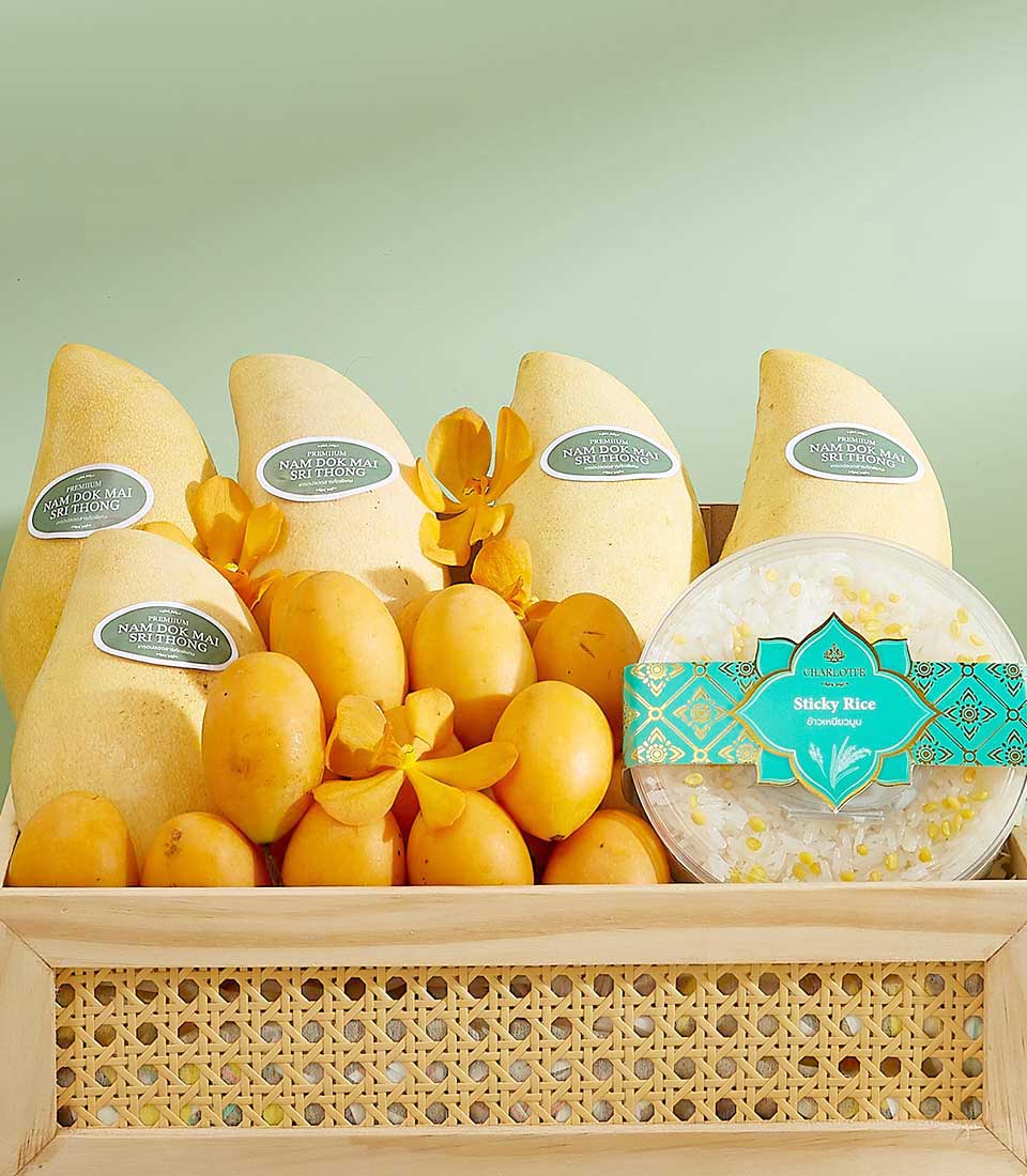 A wicker basket holds several yellow mangoes, oval-shaped mini mangoes, yellow orchids, and a box of sticky rice wrapped in turquoise packaging with a pattern. The mangoes have labels reading 