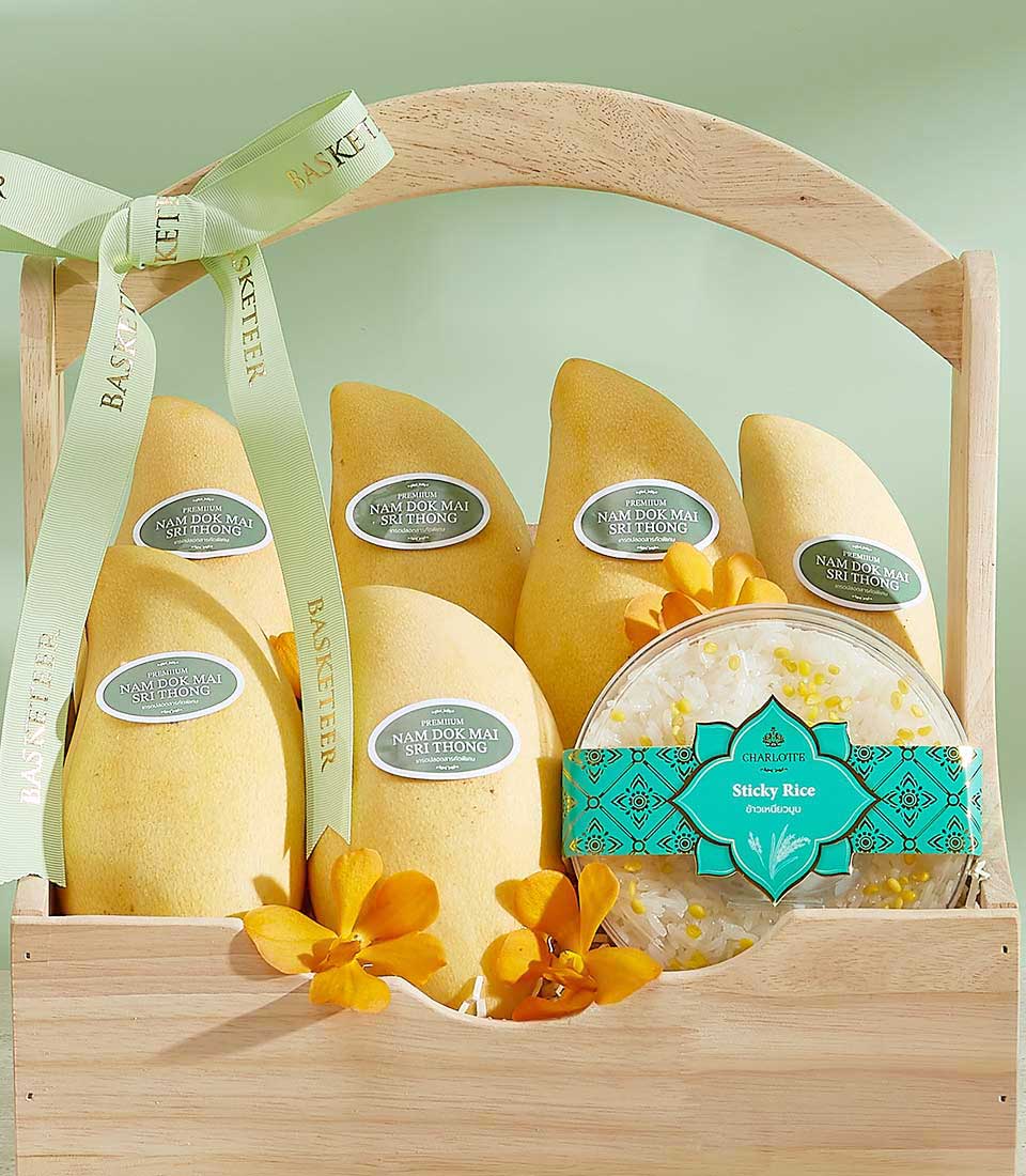 A wooden basket, dubbed the Vibrant Si Thong Mango Basket, contains six golden Nam Dok Mai mangoes with labels, a packaged sticky rice dessert in a clear container and teal wrapper, and a few yellow orchids. The basket is adorned with a light green ribbon tied in a bow with 