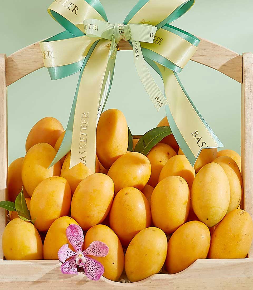 A wooden basket filled with ripe, yellow Mayongchid fruits is adorned with a green and yellow ribbon tied into a bow. A pink orchid flower is placed on top of the exotic mangoes, adding a touch of color and elegance. The background is a light green color.