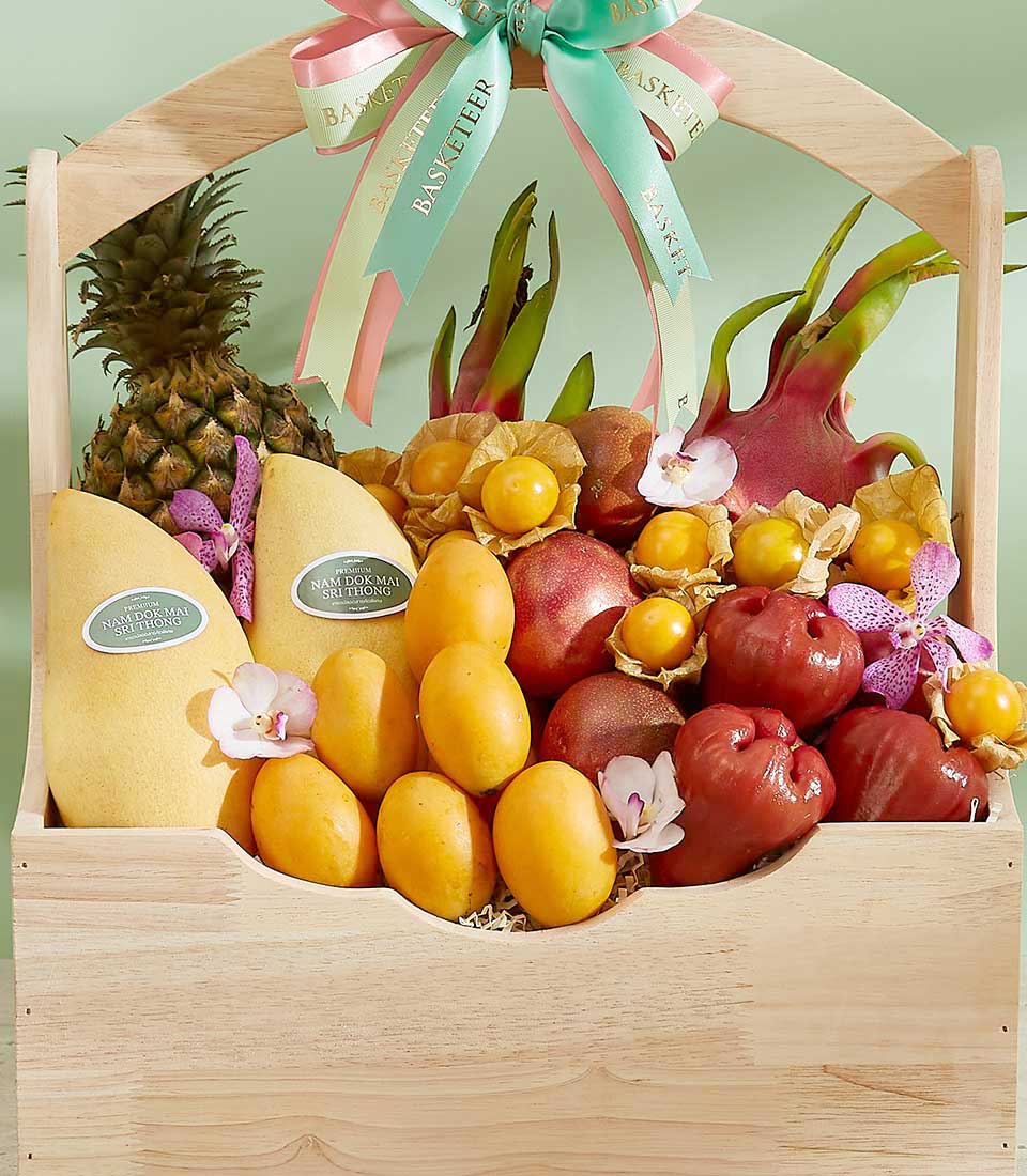 A Bountiful Thai Fruits Basket features a wooden container filled with colorful tropical delights like pineapple, dragon fruits, yellow mangoes, passion fruits, and water apples. Adorned with pink and green ribbons tied in a bow at the top and small decorative flowers, it makes for a vibrant gift.