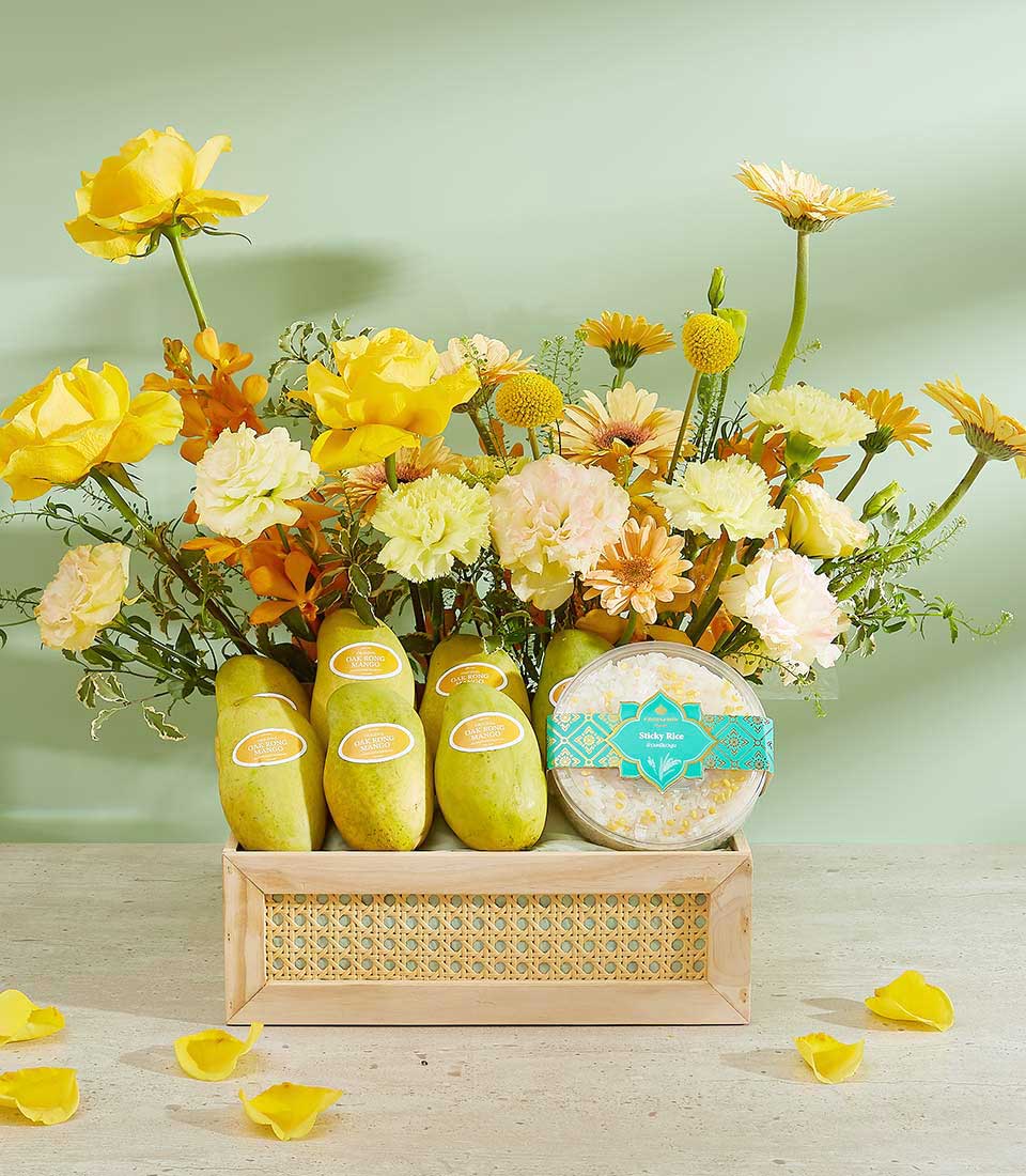 A wooden rectangular box, dubbed the Tropical Bliss Mango Basket, holds a bouquet of yellow and orange flowers, including roses and gerbera daisies, alongside four green mangoes and a round item wrapped in green and yellow packaging. Yellow petals are scattered on the table around the box.
