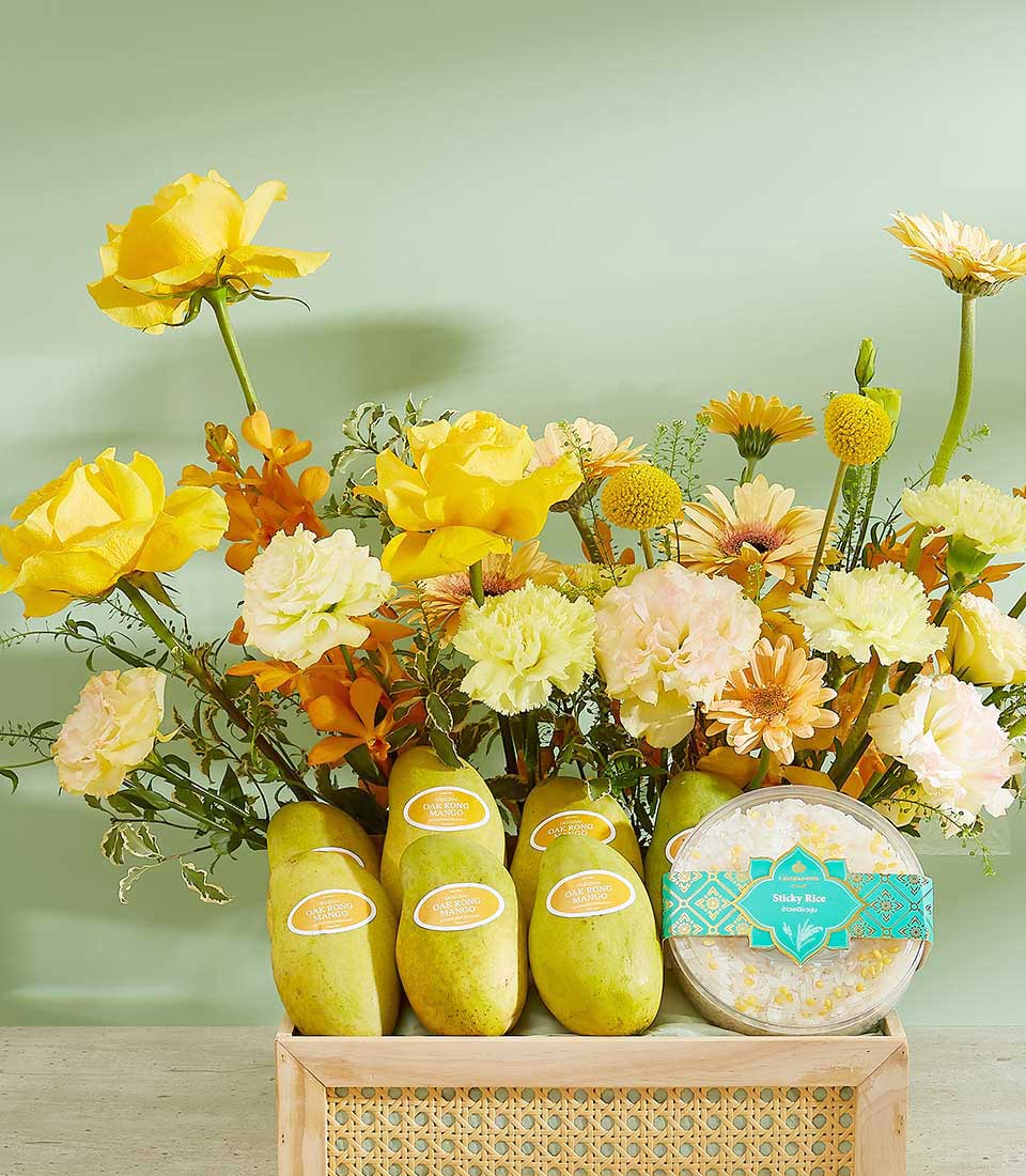 A floral arrangement of yellow and light pink flowers stands behind five labeled mangoes, lush with Tropical Bliss. They're all placed on a wooden crate beside a round container of dried flowers labeled 