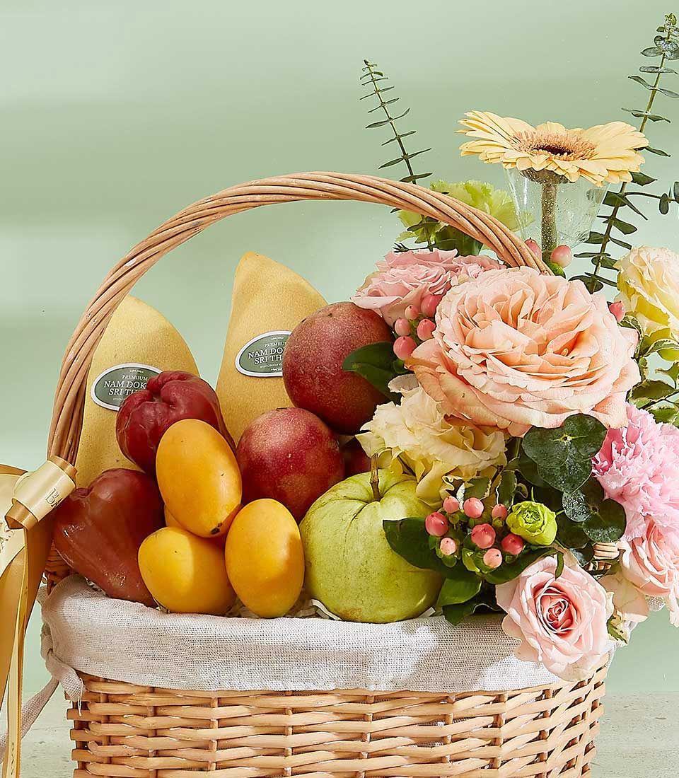 Introducing the Thai Temptations Fruit Basket: A wicker basket filled with fresh mangoes, apples, and pears, surrounded by vibrant roses and gerbera daisies amidst lush green foliage. The muted green background adds a touch of serenity to this beautiful arrangement.