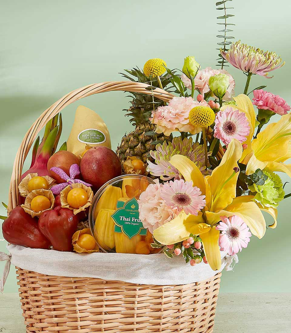 A wicker basket filled with assorted fresh fruits, including a pineapple, pears, and exotic treasures from the Thai Fruits Treasures Basket, alongside a vibrant floral arrangement with yellow lilies and pink blossoms, all set against a soft green background.